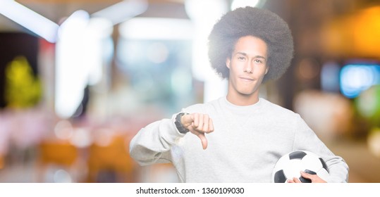 Young African American Man With Afro Hair Holding Soccer Football Ball With Angry Face, Negative Sign Showing Dislike With Thumbs Down, Rejection Concept