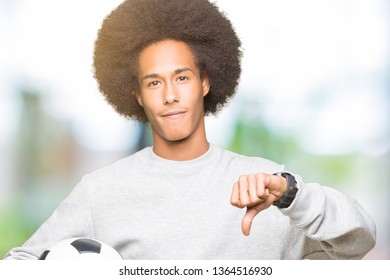 Young African American Man With Afro Hair Holding Soccer Football Ball With Angry Face, Negative Sign Showing Dislike With Thumbs Down, Rejection Concept