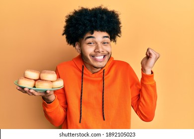 Young african american man with afro hair holding pile of tasty sugar doughnuts screaming proud, celebrating victory and success very excited with raised arm  - Shutterstock ID 1892105023