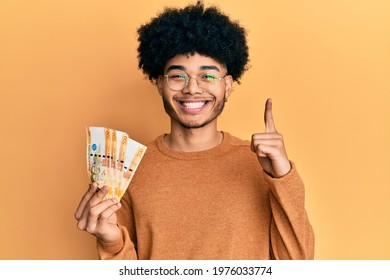 Young african american man with afro hair holding 500 philippine peso banknotes smiling with an idea or question pointing finger with happy face, number one 