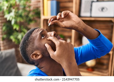 Young African American Man Applying Eye Drop Sitting On Sofa At Home