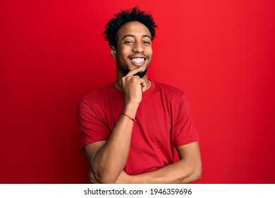 Young african american man with beard wearing casual red t shirt looking confident at the camera with smile with crossed arms and hand raised on chin. thinking positive.  - Powered by Shutterstock