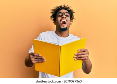 Young African American Man With Beard Reading A Book Wearing Glasses Smiling And Laughing Hard Out Loud Because Funny Crazy Joke. 