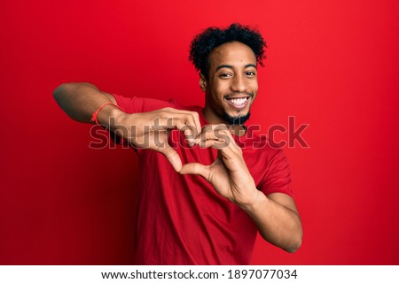 Young african american man with beard wearing casual red t shirt smiling in love doing heart symbol shape with hands. romantic concept. 