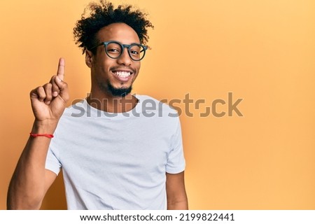 Young african american man with beard wearing casual white t shirt and glasses smiling with an idea or question pointing finger up with happy face, number one 