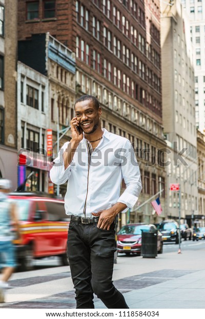 Young African American Man with beard\
traveling in New York, wearing white shirt, black pants, walking on\
street in Manhattan, talking on cell phone, smiling. Cars,\
buildings on\
background.\
