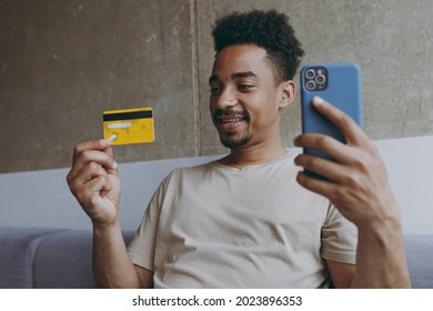 Young African American Man In Beige T-shirt Sitting On Grey Sofa Indoors Apartment Hold Mobile Cell Phone Credit Bank Card Shopping Online Order Delivery Booking Tour Rest On Weekends Stay At Home.