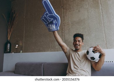 Young African American Man In Beige T-shirt Cheer Up Support Favorite Team With Soccer Ball Football Fan Foam Glove Finger Up Scream Sit On Grey Sofa Indoors Apartment People Sport Leisure Concept.