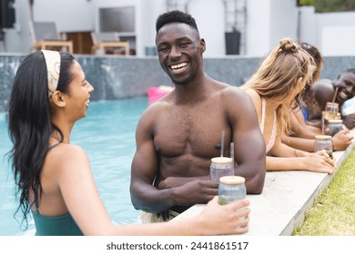 A young African American man with a bright smile is enjoying a poolside conversation. He is accompanied by a diverse group of friends, sharing drinks and laughter in a relaxed outdoor setting. - Powered by Shutterstock