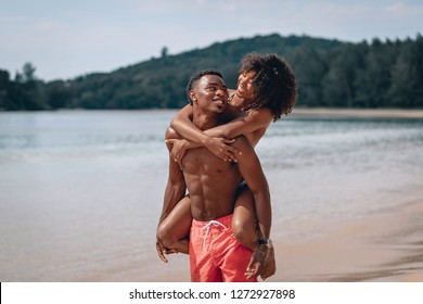 Young African american man carrying his smiling girlfriend on his back while enjoying a late afternoon together at the beach. Phuket. Thailand.