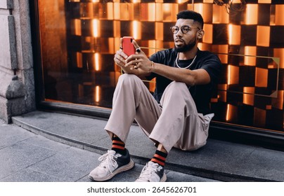 Young African American man in casual outfit sitting on urban sidewalk, holding smartphone with focused expression, possibly checking mobile banking app or trading financial data