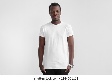 Young African American Man With Dark Skin Standing With Hands In Pockets, Wearing Blank White T Shirt With Copy Space For Your Logo Or Text, Isolated On Grey Background