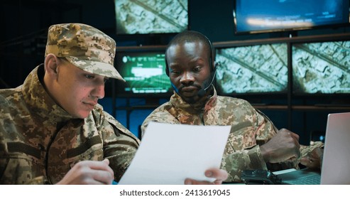 Young African American man dispatcher in headset working on laptop computer in control room. Multiethnic soldiers sitting at table and studying secret documents of military operation and strategy. - Powered by Shutterstock