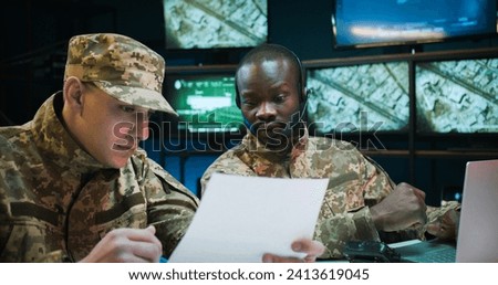 Young African American man dispatcher in headset working on laptop computer in control room. Multiethnic soldiers sitting at table and studying secret documents of military operation and strategy.
