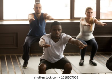 Young African American Man Doing Squat Exercise At Group Fitness Training, Sporty Black Guy Focused On Self-improvement Working Out With Diverse Active People In Gym Studio During Routine Session