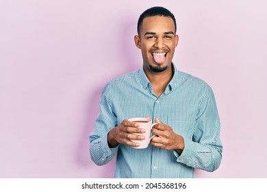 Young African American Man Drinking A Cup Of Coffee Sticking Tongue Out Happy With Funny Expression. 