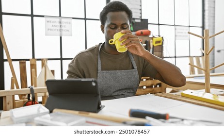 A young african american man drinks coffee while using a tablet in a woodwork workshop. - Powered by Shutterstock