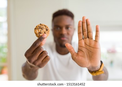 Young african american man eating chocolate chips cookies with open hand doing stop sign with serious and confident expression, defense gesture
