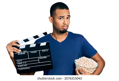 Young African American Man Eating Popcorn Holding Film Clapboard Clueless And Confused Expression. Doubt Concept. 