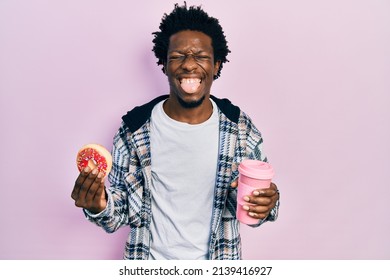 Young african american man eating doughnut and drinking coffee sticking tongue out happy with funny expression. 