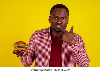 Young African American Man Eating Hamburger Isolated On Yellow Background.real Jam Licking Fingers