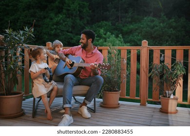 Young African american man enjoys playing the guitar with little children accompaining him with instruments on patio in garden in summer. - Powered by Shutterstock