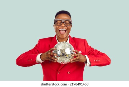 Young African American Man Entertainer In Stylish Glasses And Red Suit Clutches Disco Ball Symbolizing Relaxation In Nightclub And Fun Rave Standing On Pastel Blue Background. Party Producer Portrait