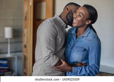 Young African American man giving his laughing wife a kiss on the cheek in their living room at home