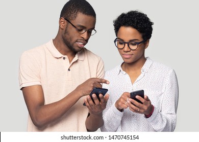 Young african american man in glasses showing new mobile software app to smiling wife. Happy black family couple sharing photos or contacts, using smartphones, isolated on grey studio background.
