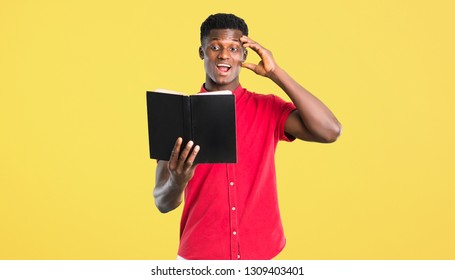 Young African American Man Holding A Book And Surprised While Enjoying Reading On Yellow Background