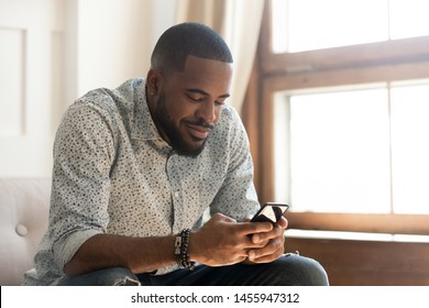 Young african american man holding smartphone texting message or play mobile game sit on sofa at home, smiling millennial black guy using social media app messenger, surfing web on phone indoors