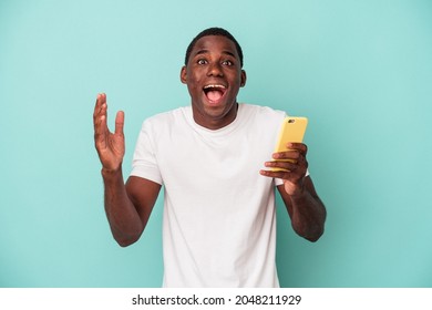 Young African American Man Holding A Mobile Phone Isolated On Blue Background Receiving A Pleasant Surprise, Excited And Raising Hands.