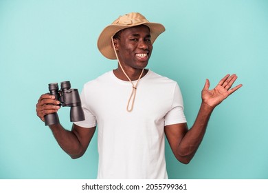 Young African American Man Holding Binoculars Isolated On Blue Background Showing A Copy Space On A Palm And Holding Another Hand On Waist.