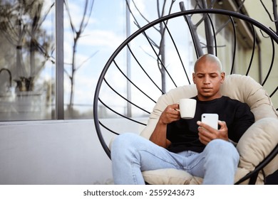 Young african american man holding coffee and using smartphone on armchair on balcony	 - Powered by Shutterstock