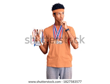 Young african american man holding champion trophy wearing medals serious face thinking about question with hand on chin, thoughtful about confusing idea 