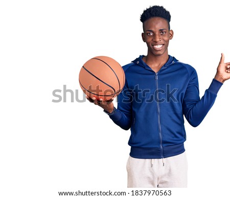 Young african american man holding basketball ball smiling happy pointing with hand and finger to the side 