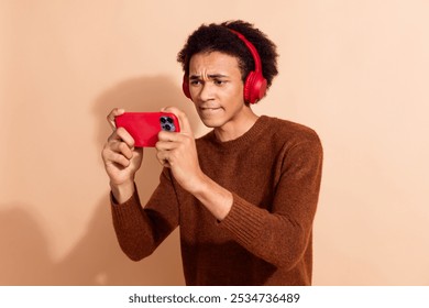 Young african american man intensely playing mobile game with red phone and headphones in casual outfit against beige background - Powered by Shutterstock