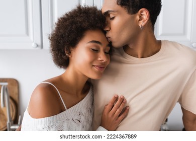 Young African American Man Kissing Smiling Girlfriend Standing With Closed Eyes In Kitchen