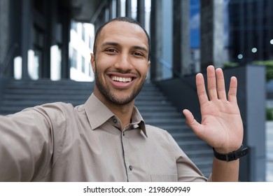 Young African American Man Looking Into Smartphone Camera, Talking On Video Call Using App On Phone, Smiling And Happy Waving Hand Gesture Of Greeting, Businessman Outside Modern Office Building.