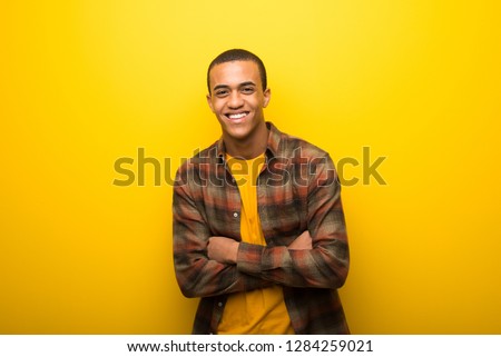 Young african american man on vibrant yellow background keeping the arms crossed while smiling