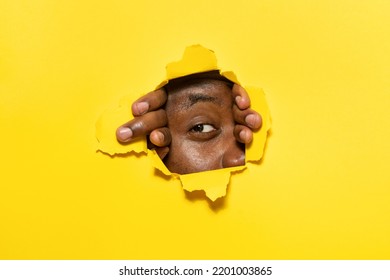 Young African American Man Peeking Out Of A Hole In Yellow Paper Background, Look Through Torn Hole With One Eye, Free Copy Space