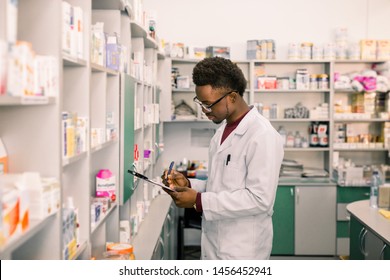 Young African American man pharmacist standing in interior of pharmacy. Man specialist of pharmacy making notes on clipboard during inventory