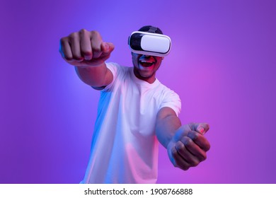 Young african american man playing video games in VR-glasses, excited black millennial guy experiencing virtual reality while standing in neon light over purple gradient background, copy space