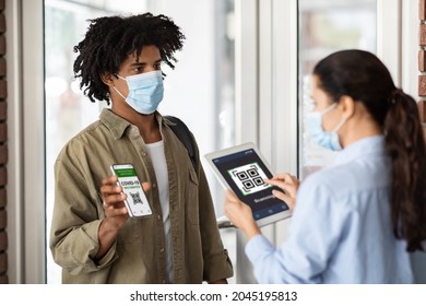 Young African American Man Showing Vaccination Certificate On Smartphone At Cafe Entrance, Female Cafeteria Worker In Medical Mask Using Digital Tablet For Screening Health Qr Code Of Customer