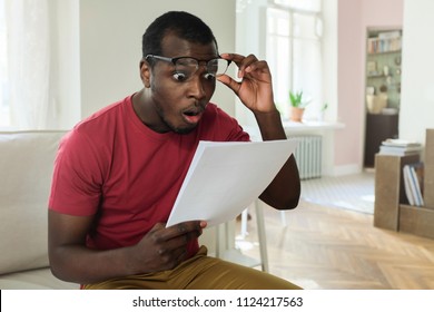 Young african american man sitting on couch in modern apartment with mouth open, holding utility bill with high rates, raised eyeglasses in wow or surprise gesture 