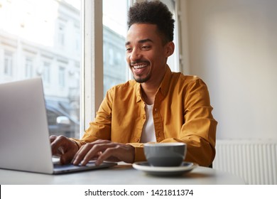 Young African American man sitting at a table in a cafe and working on a laptop, wears yellow shirt, drinks aromatic coffee, creates new content for his blog, enjoys the work of a freelancer.