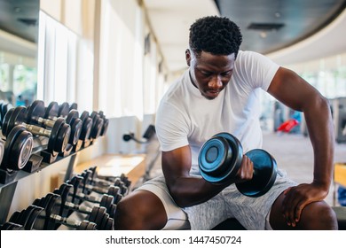 Young African American man sitting and lifting a dumbbell close to the rack at gym. Male weight training person doing a biceps curl in fitness center