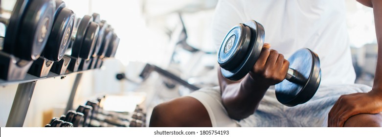 Young African American man sitting and lifting a dumbbell close to the rack at gym. Male weight training person doing a biceps curl in fitness center - Powered by Shutterstock