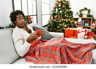 Young African American Man Sitting On The Sofa Drinking Coffee By Christmas Tree Laughing And Embarrassed Giggle Covering Mouth With Hands, Gossip And Scandal Concept 