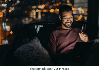 Young African AMerican man sitting on balcony with urban view and using tablet at night - Powered by Shutterstock